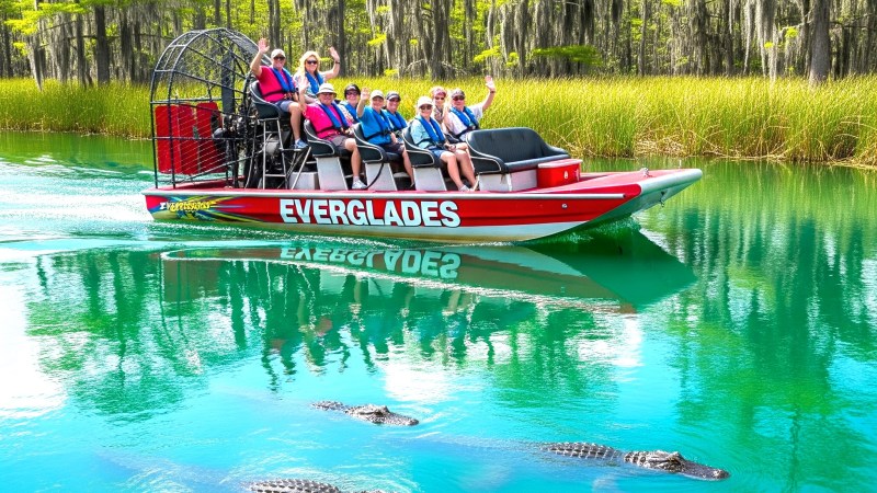 Tourists on an airboat in the Everglades with alligators visible in the water.